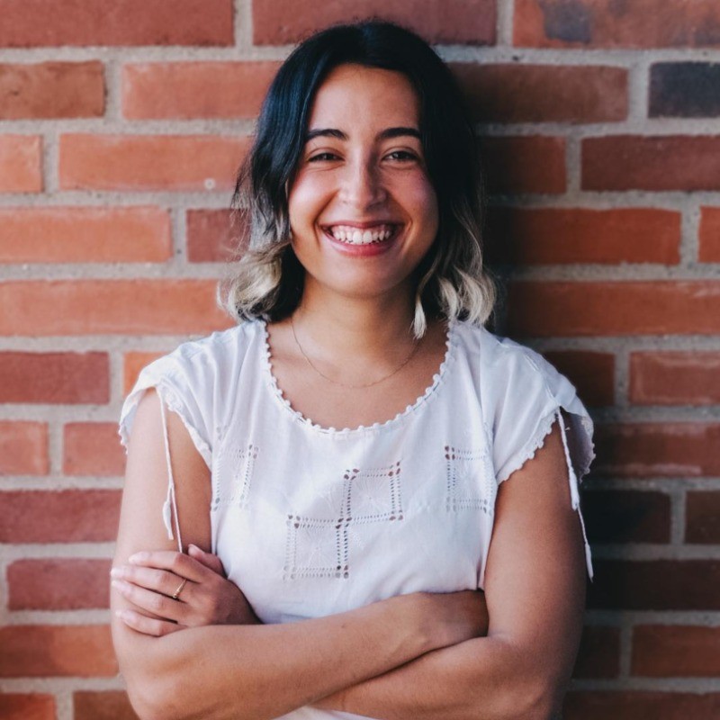 A smiling woman with shoulder-length dark hair with light tips, wearing a white blouse, standing with arms crossed in front of a brick wall.